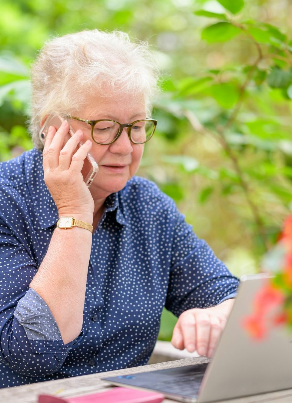 Seniorin am Telefon Ältere Frau mit Brille, Telefon am Ohr, arbeitet am Laptop in einer grünen Umgebung.